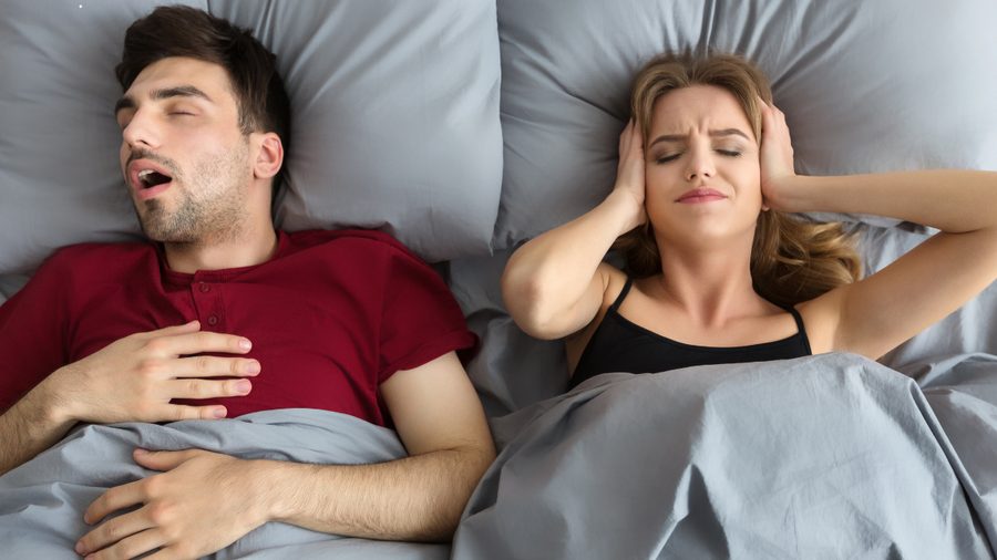Una pareja en la cama: él duerme boca arriba roncando con la boca abierta y ella se tapa los oídos con las manos